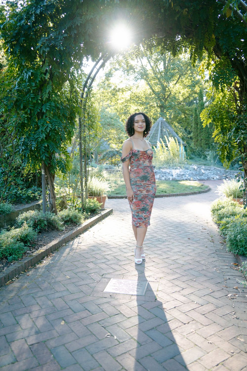 Young woman in floral midi dress walking under green garden archway in bright sunlight.