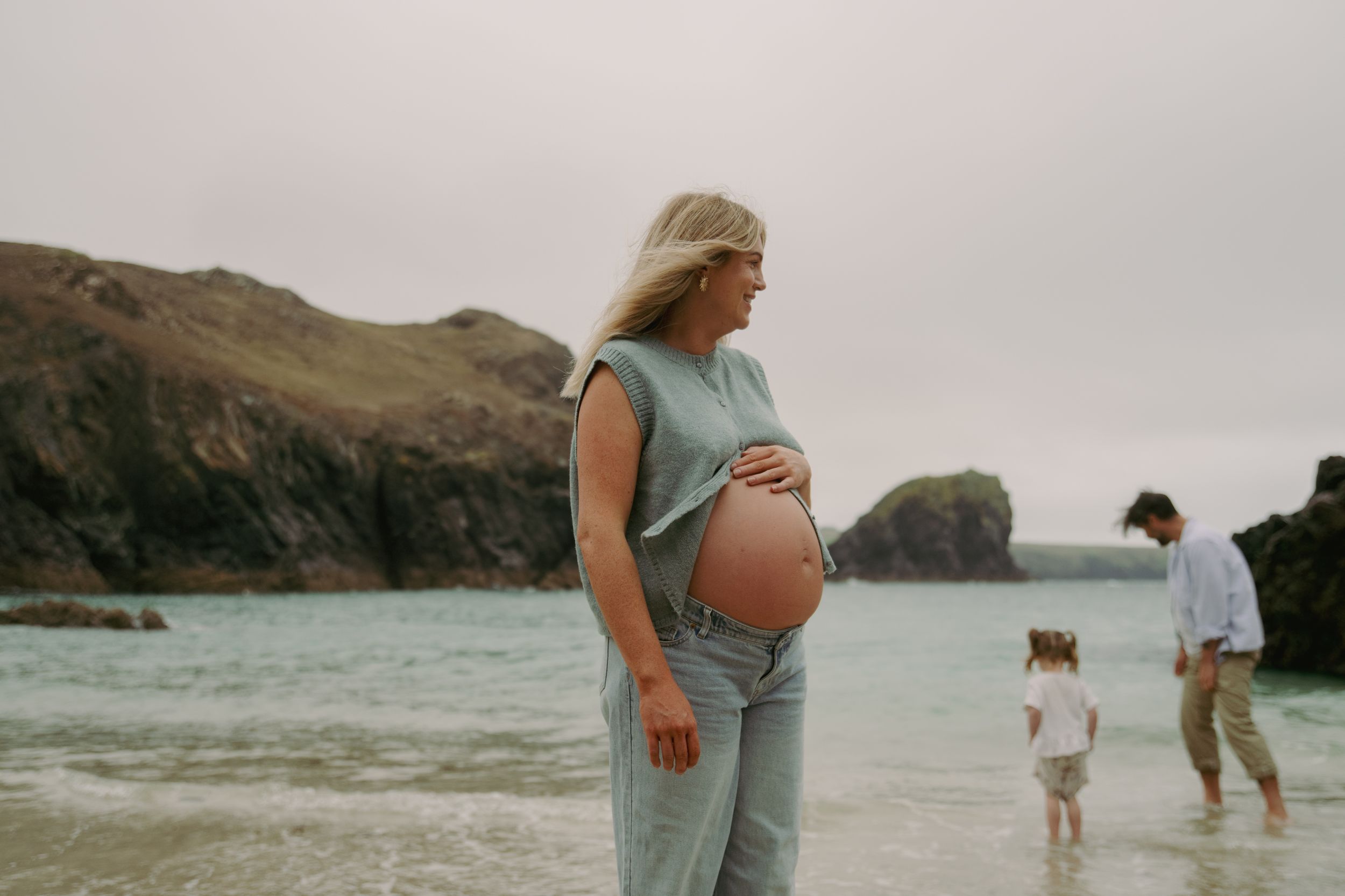 A maternity photo series captures intimate moments of a couple at a misty beach with mountains in the background.