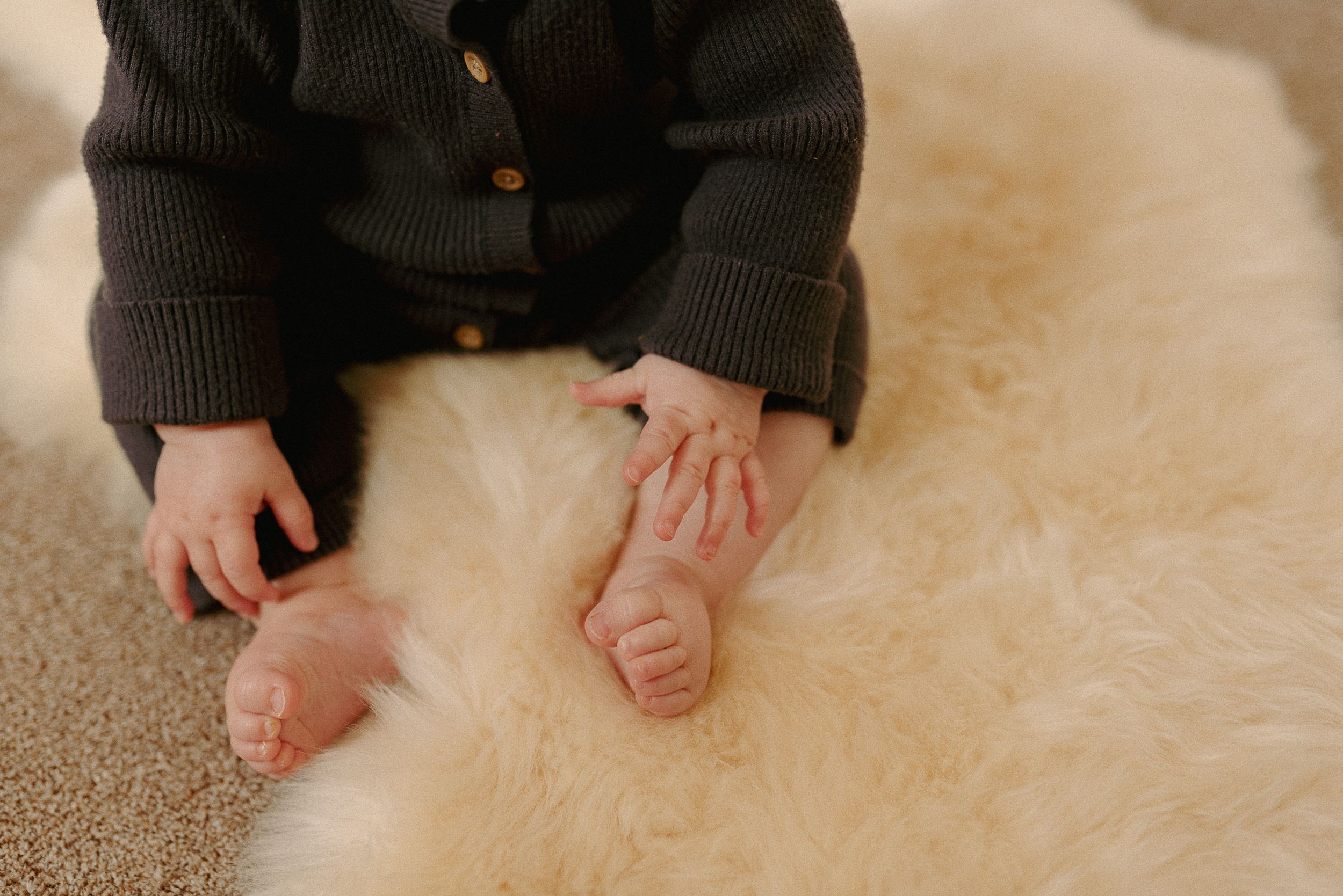 Tiny bare feet and hands rest gently on soft beige carpet next to a black sweater.