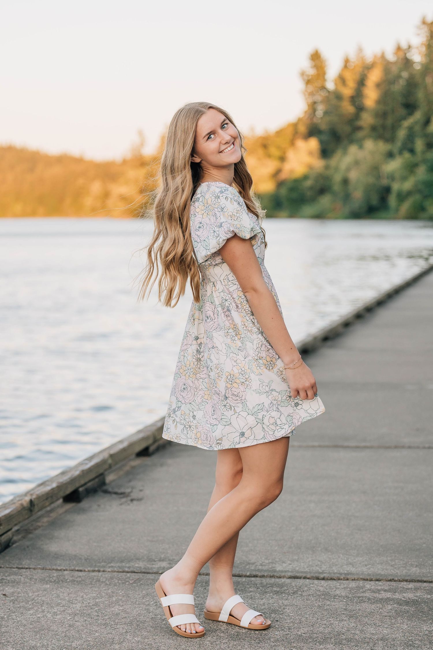 Smiling blonde girl in floral mini dress twirls on lakeside dock at sunset.