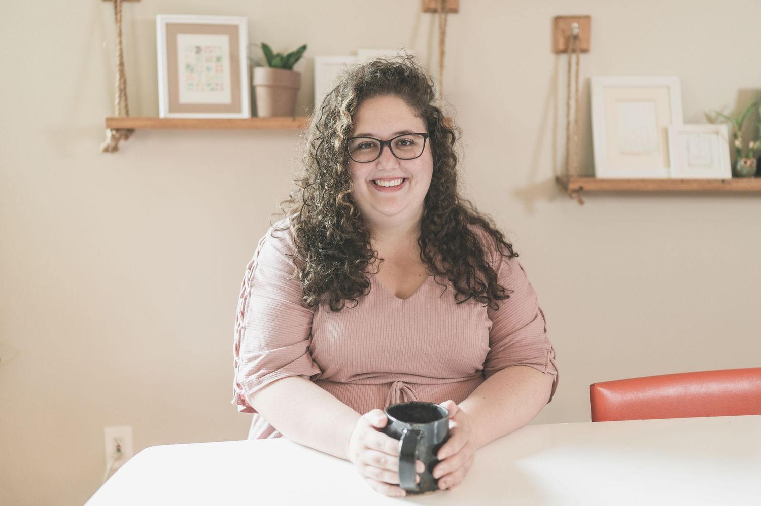 Casual indoor portrait of someone in pink holding coffee on couch with shelving in background.