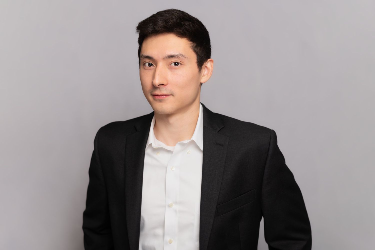 Professional headshot of someone in a black suit jacket and white shirt against a gray background.