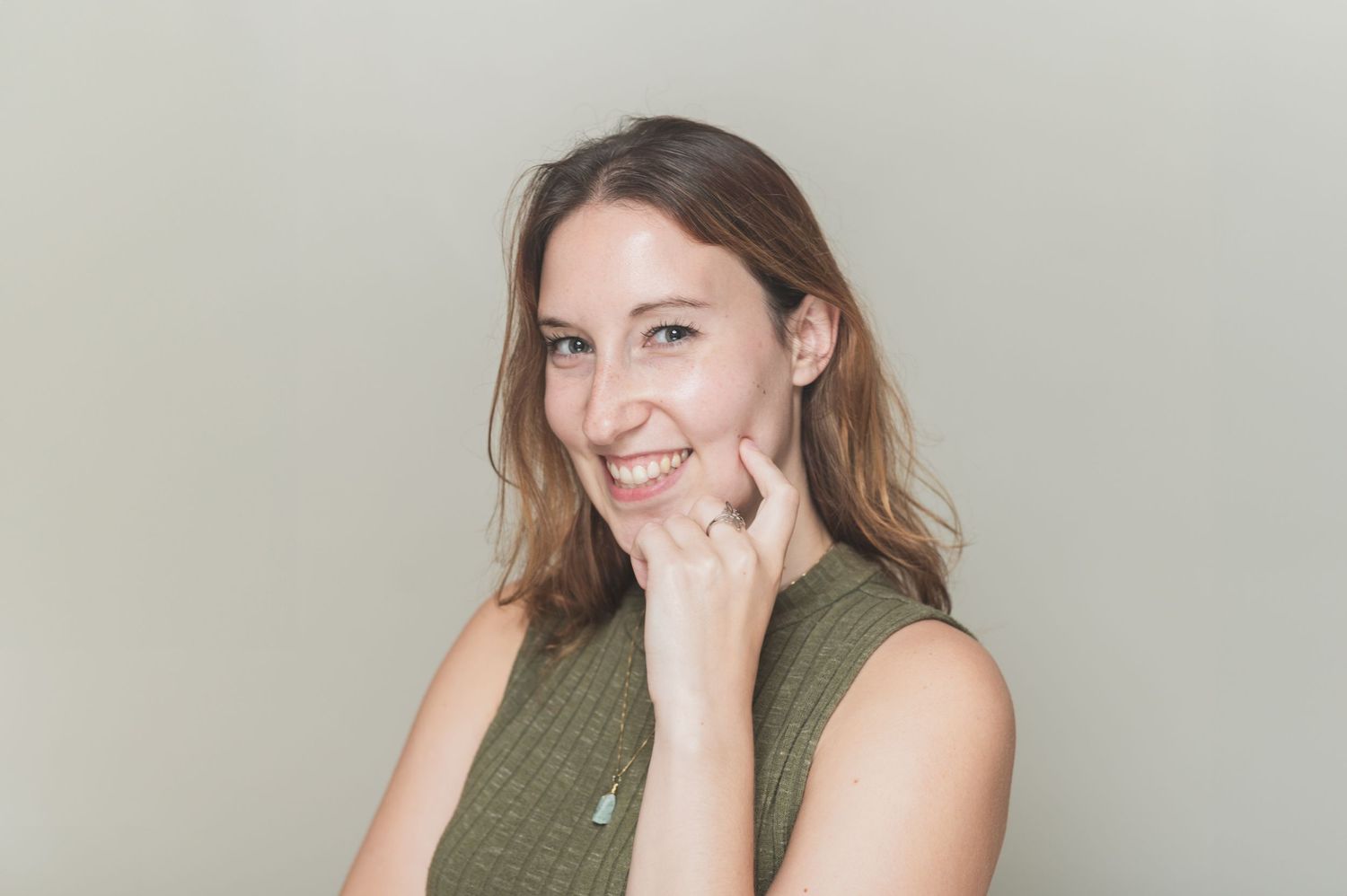 Smiling person with long brown hair wearing an olive green sleeveless top poses against neutral background.