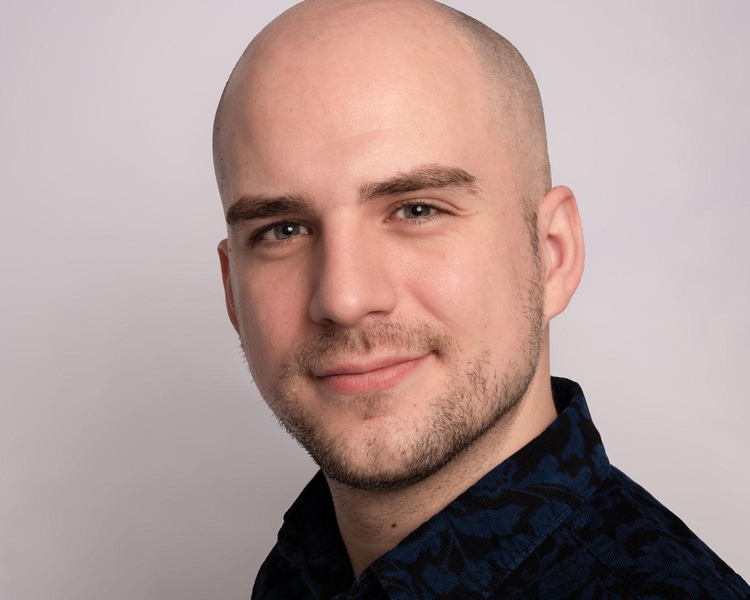 A professional headshot of a person wearing a dark patterned shirt against a light background.