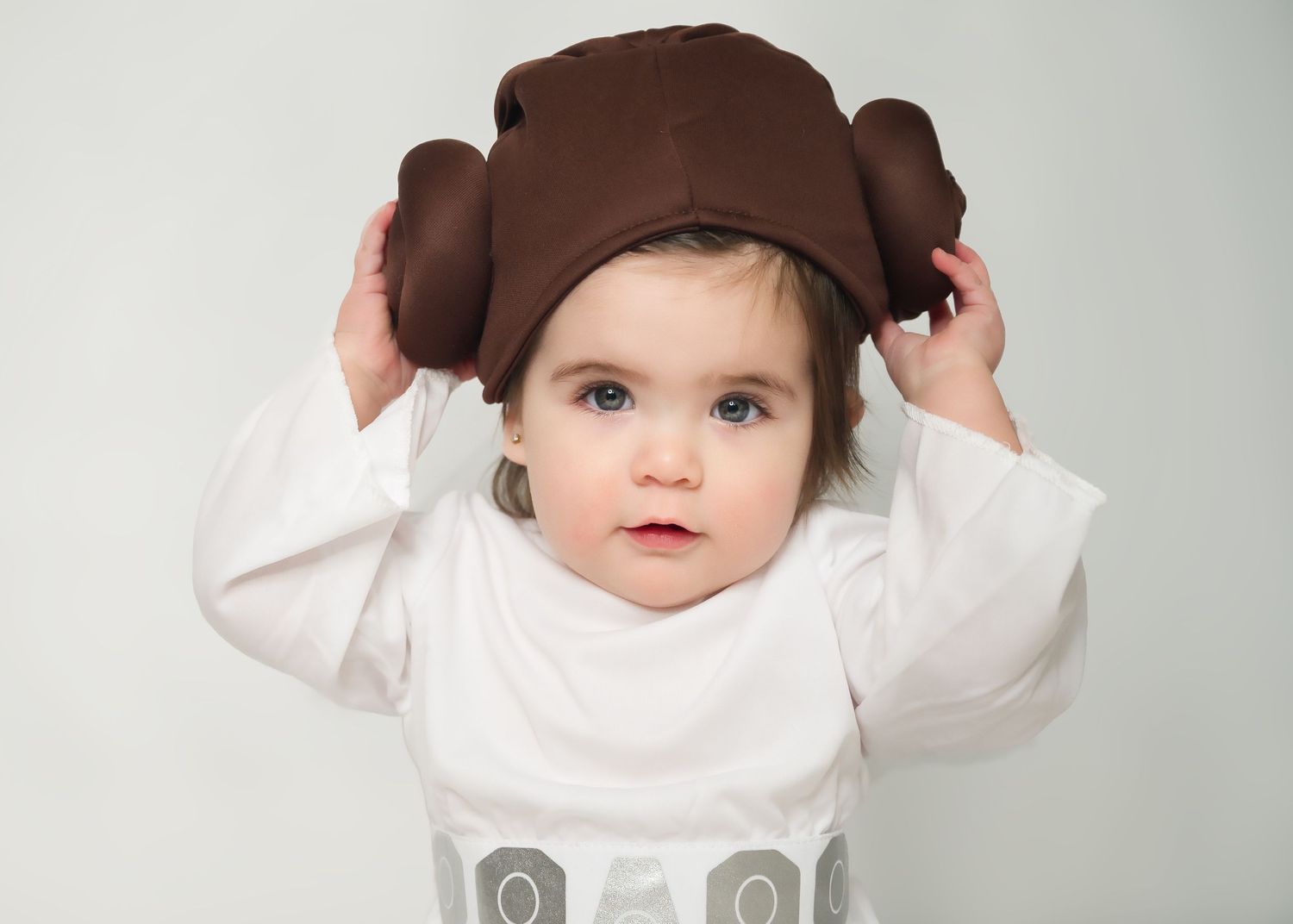 A toddler wearing a brown bear hat and white outfit looking at the camera.