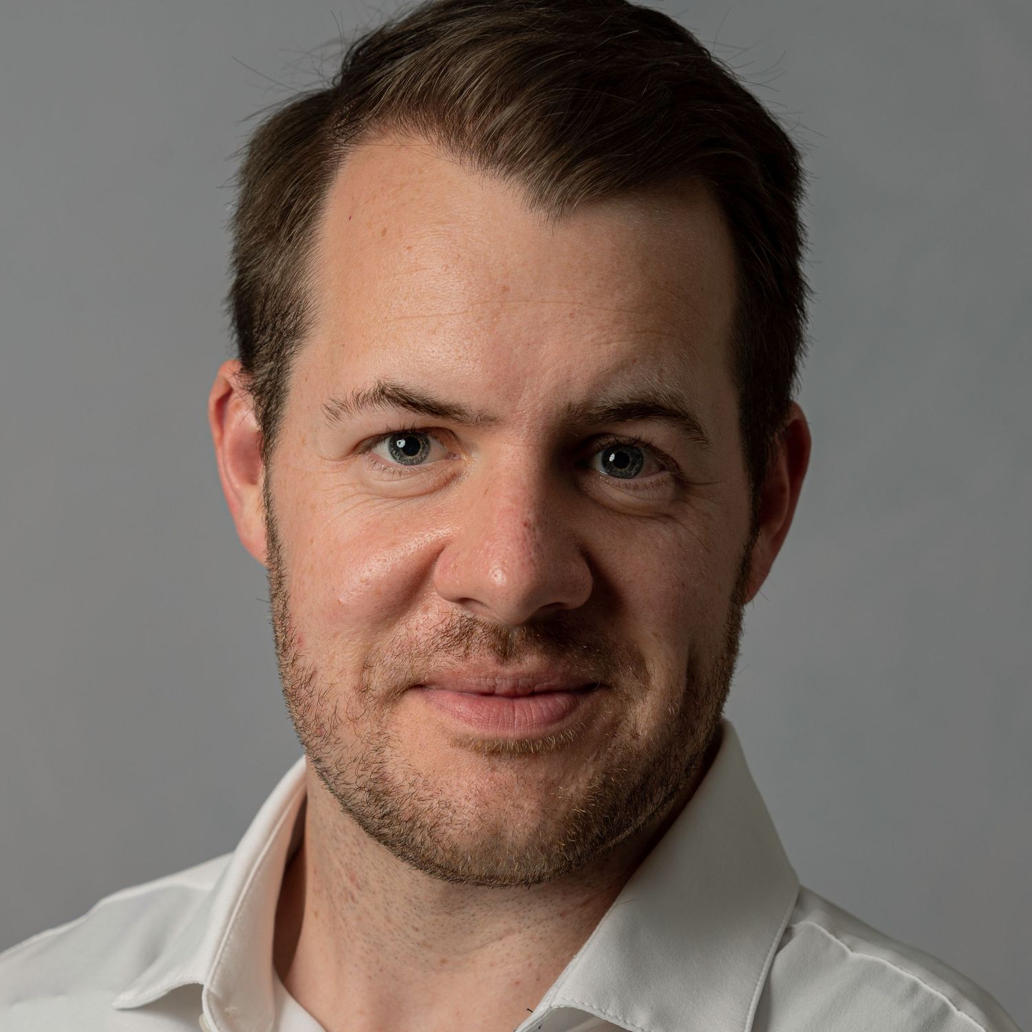 Professional headshot portrait of a man in a white dress shirt smiling against a gray backdrop.