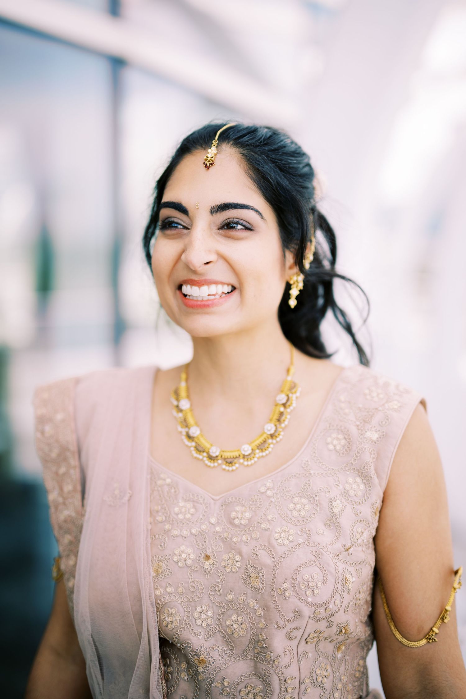 South Asian bride wearing pink embroidered dress with gold necklace and traditional bindi decoration smiles warmly.