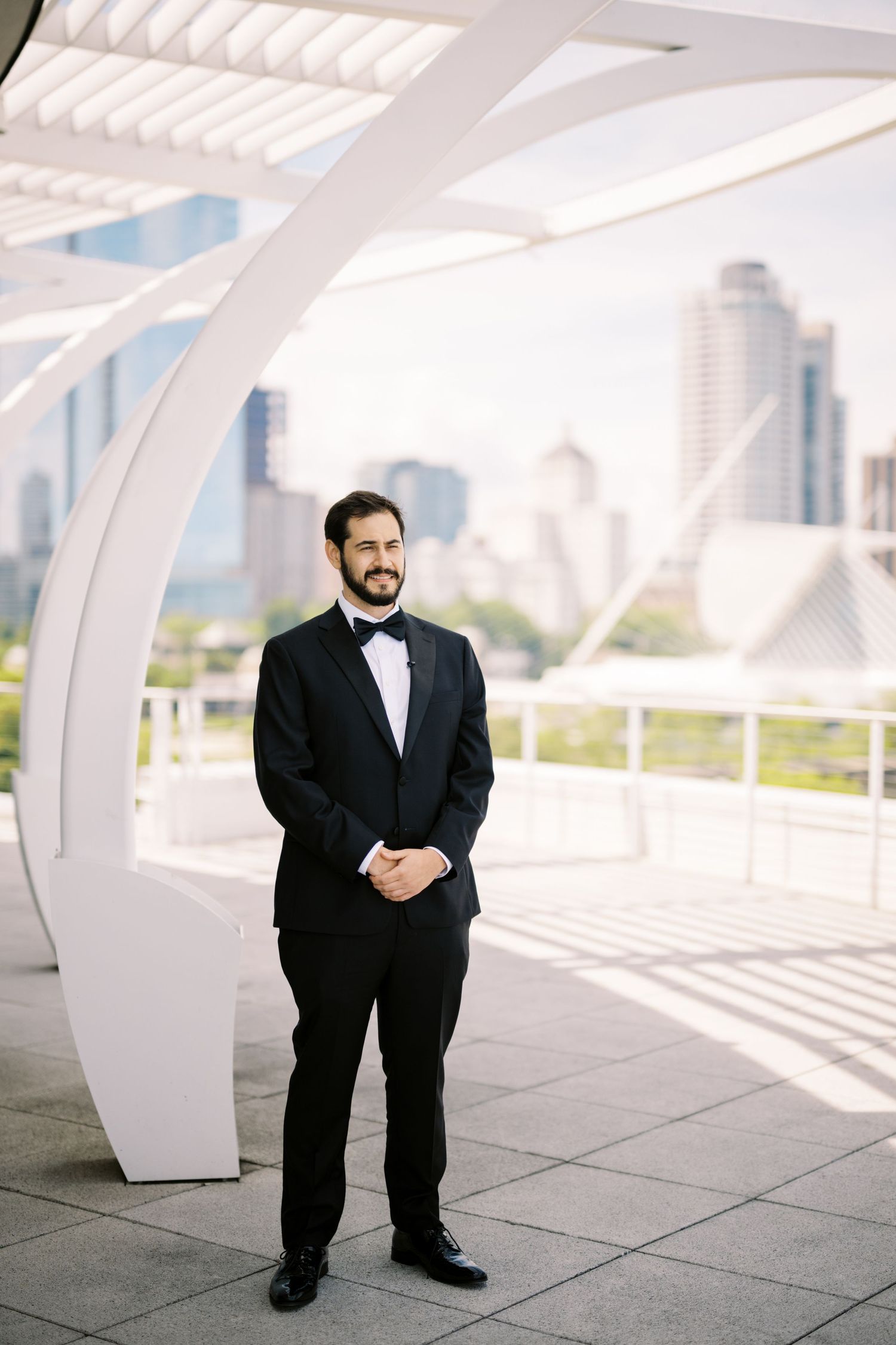 A person in a classic black tuxedo and bowtie stands in front of modern architectural curves and city skyline.