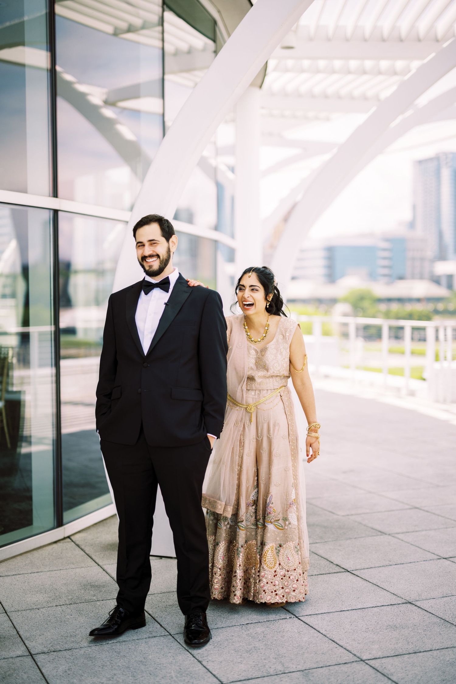A couple in formal attire stand together outdoors, with the bride wearing an elegant champagne-colored embellished gown.