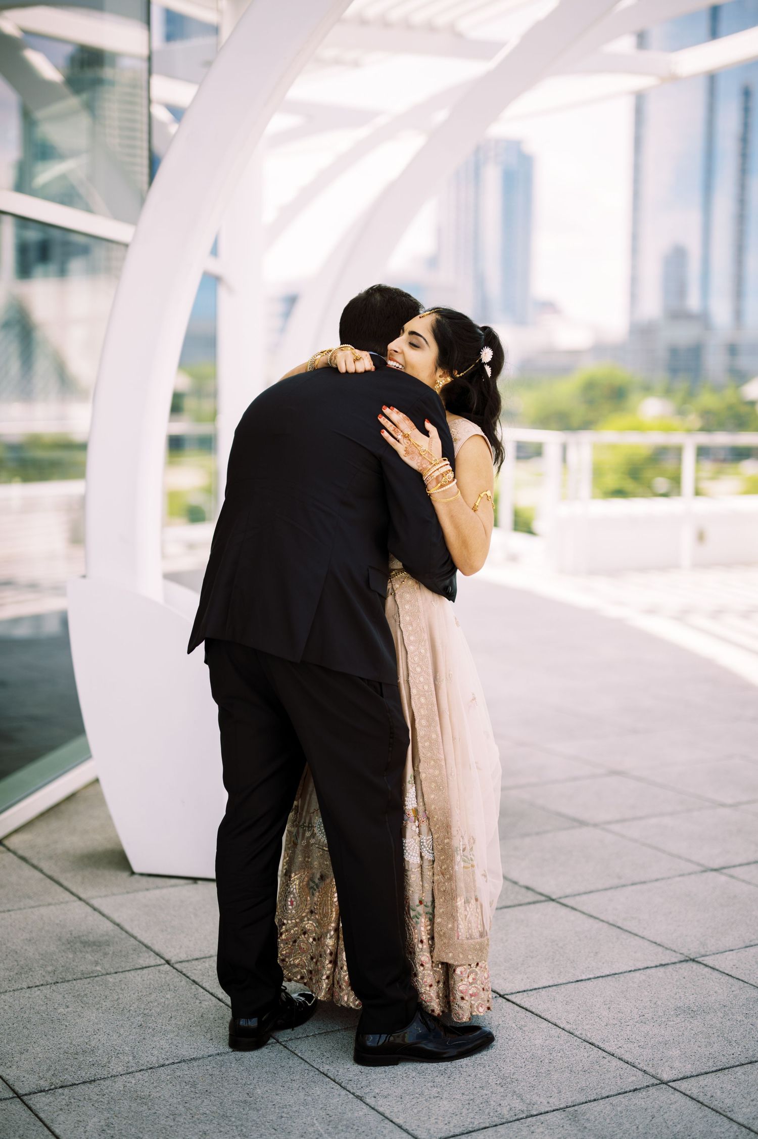 A couple embraces in an intimate moment under a modern white architectural structure with city buildings in the background.