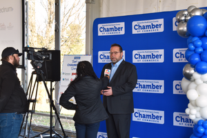 Person being interviewed by news media against a backdrop with Chamber logos and blue balloons.