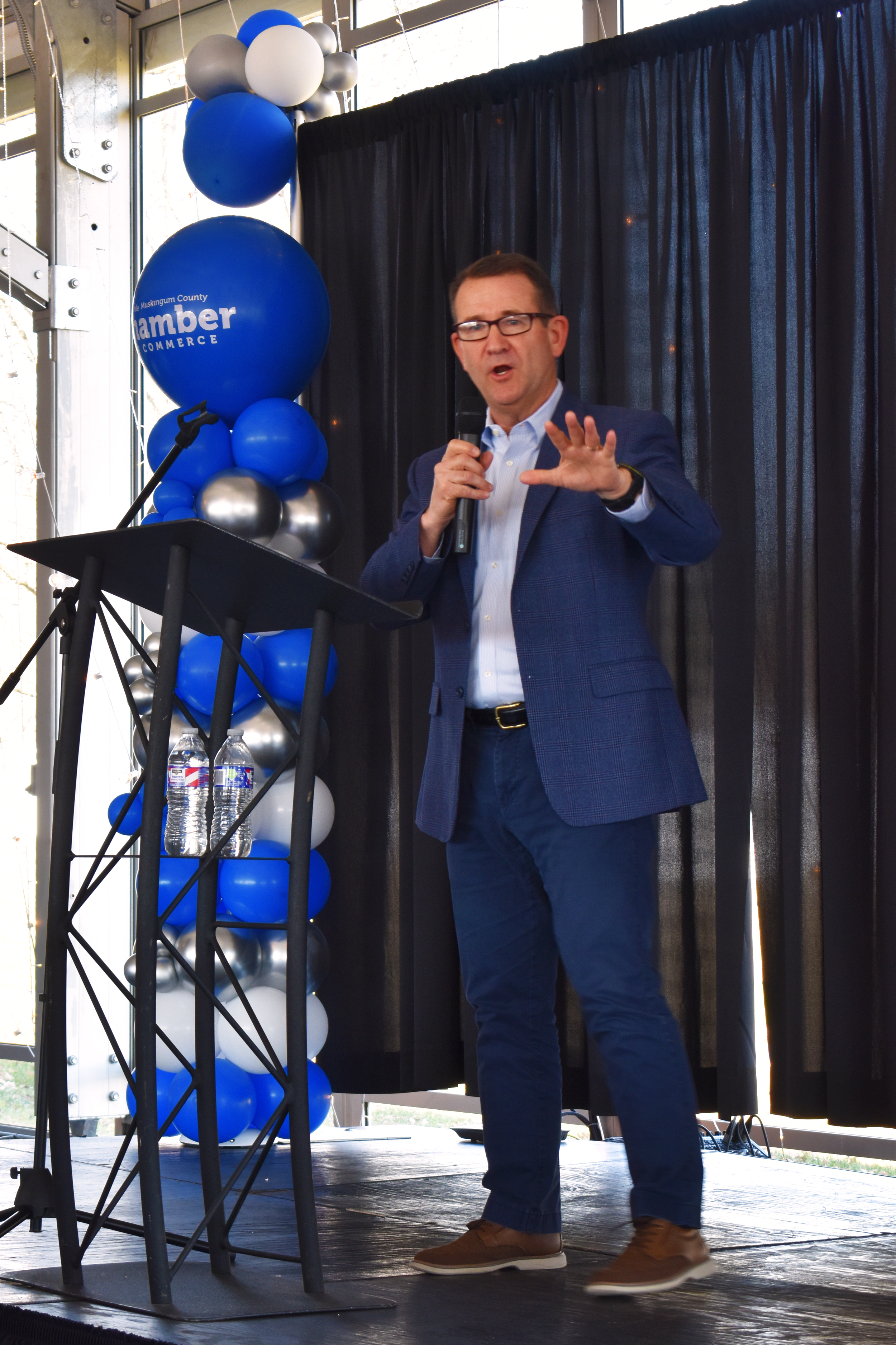 A speaker in a navy blue suit gives a presentation on stage with blue and silver balloons decorating the backdrop.