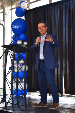 A speaker in a navy blue suit gives a presentation on stage with blue and silver balloons decorating the backdrop.