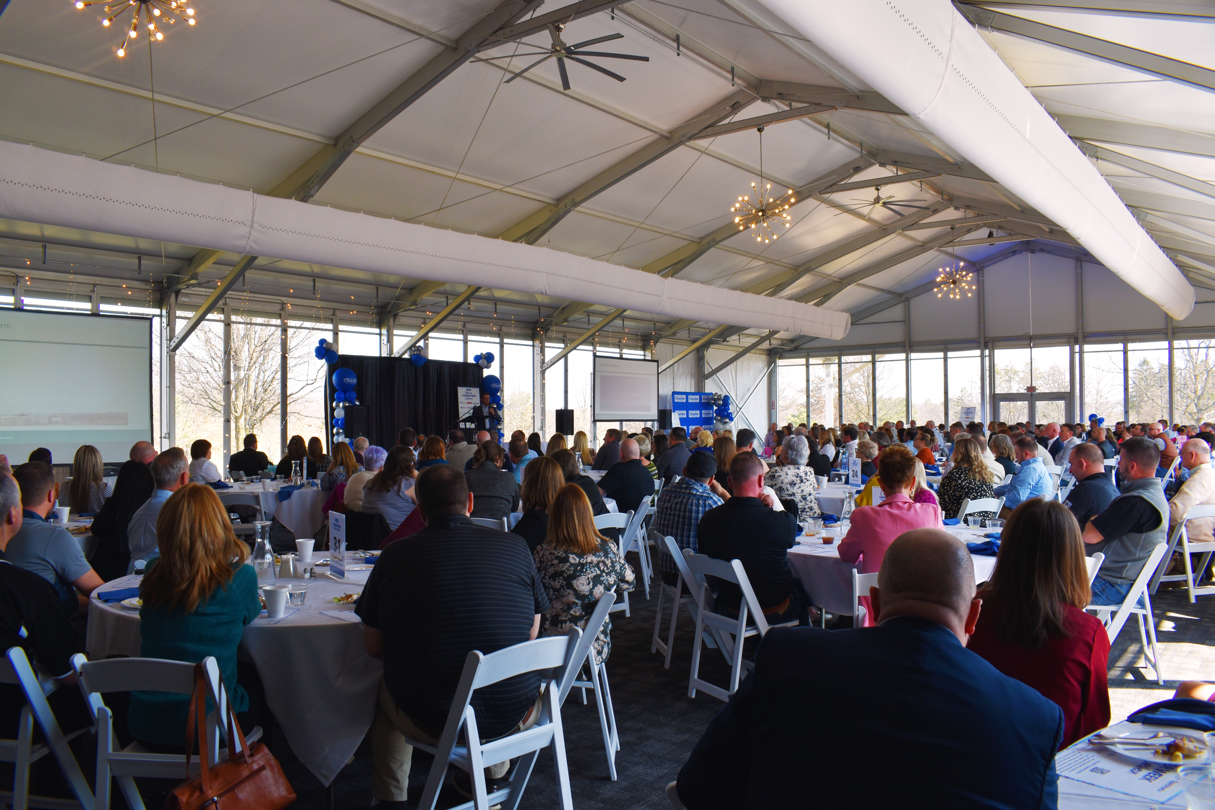 Large group of attendees seated at round tables in a brightly lit event space with white ceiling and chandeliers.