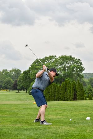 A golfer in navy shorts and gray shirt takes a backswing on a lush golf course with trees in the background.