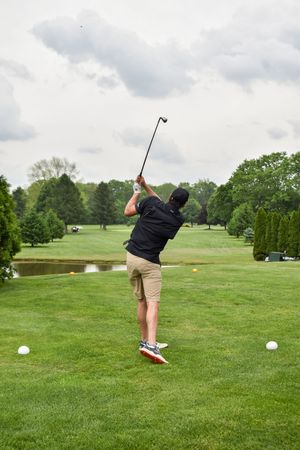 A golfer in black shirt and khaki shorts takes a swing on a lush green course with trees and a pond in the background.
