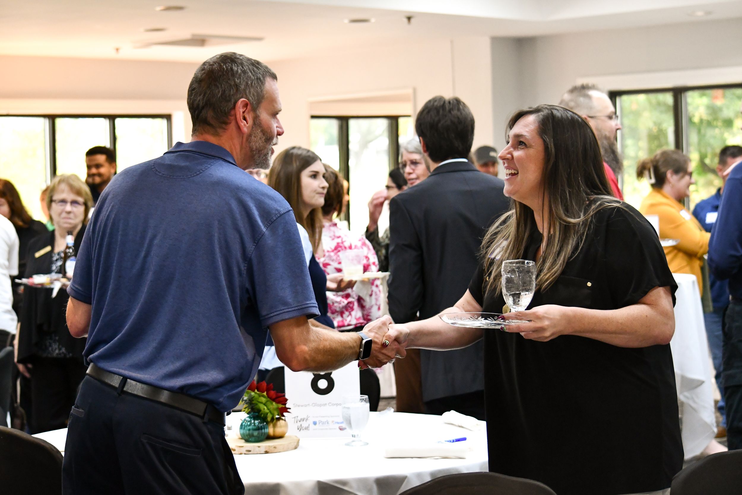Two professionals shaking hands at a networking event in a bright conference room with other attendees in background.