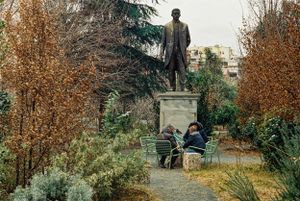 Bronze statue stands on pedestal in park with autumn trees and benches. Tirana, Albania,