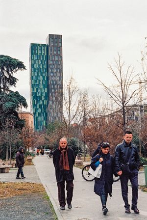 People walking on path with modern blue glass tower visible through winter trees. Tirana, Albania,