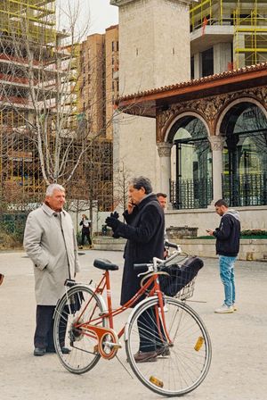 Two people chat next to an orange bicycle outside a historic building with arched windows. Tirana, Albania,