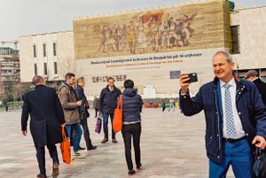 People gather in a public square with a large mural visible on the building wall. Tirana, Albania,