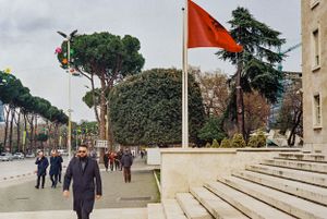 Red flag waves on a pole along a tree-lined walkway with people strolling. Tirana, Albania,