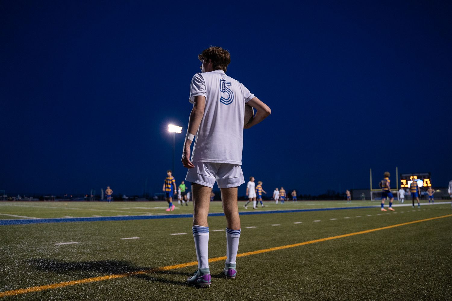 Player in white uniform stands on soccer field at night under stadium lights.