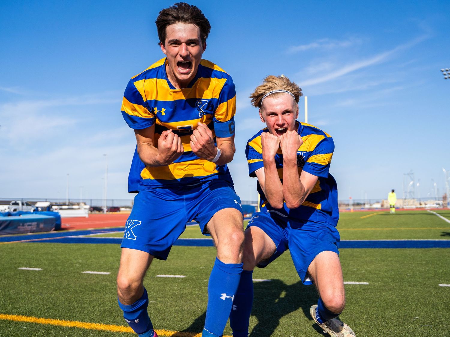 Two athletes in blue and yellow striped uniforms pose playfully on sports field.