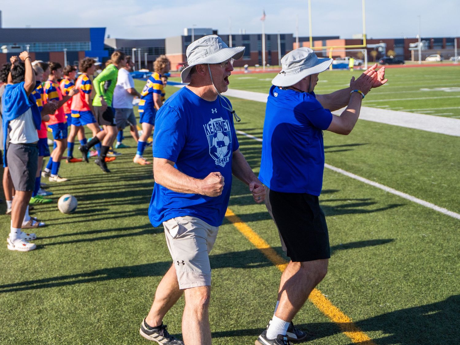 Coaches in blue shirts observe from sideline during outdoor sports event.