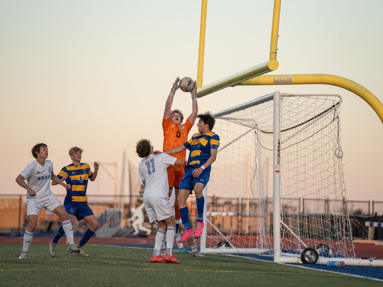 Soccer players compete for the ball near the goal post during a sunset match on an outdoor field.