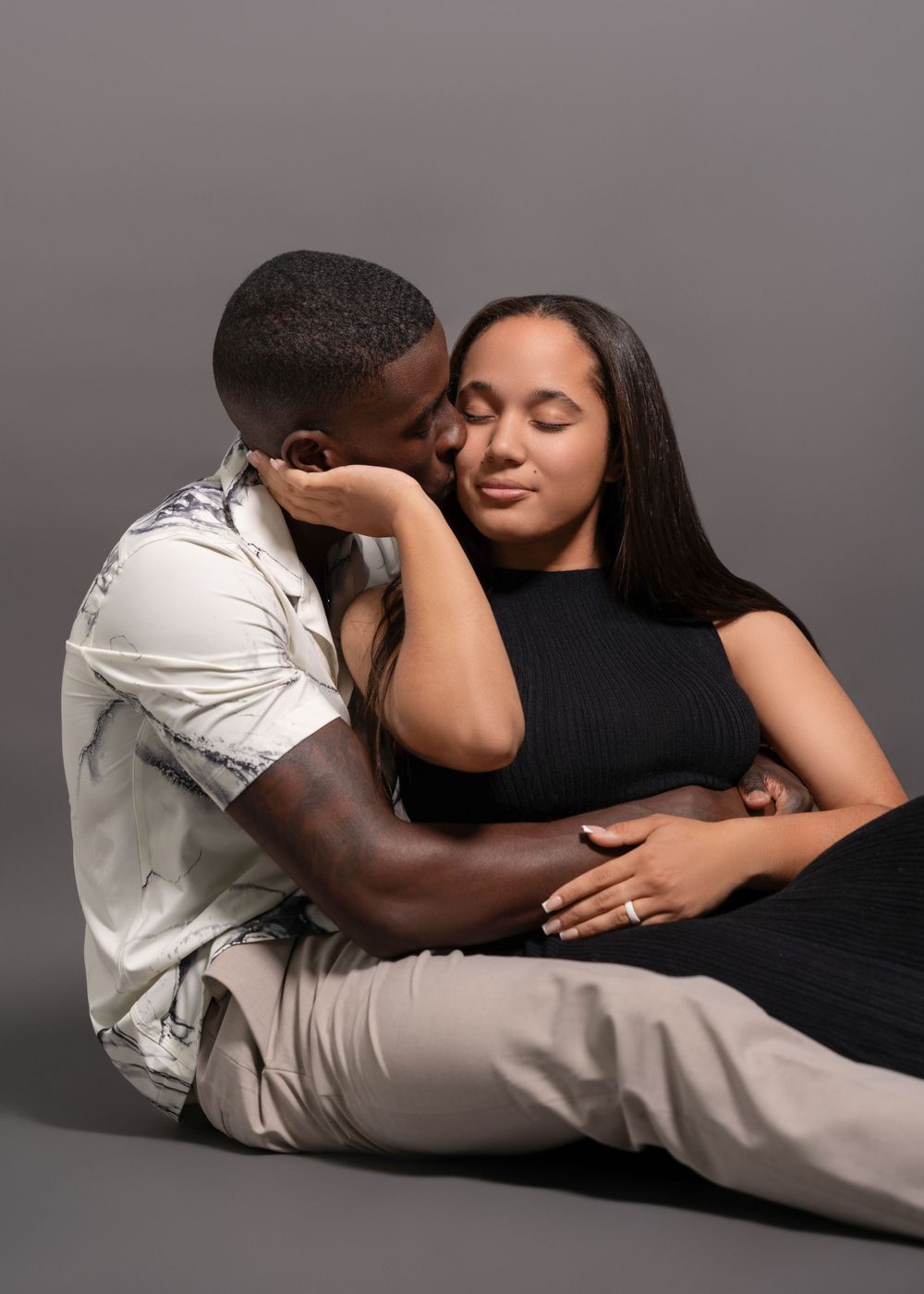 Intimate couple sharing a tender moment while seated on the floor against grey backdrop.