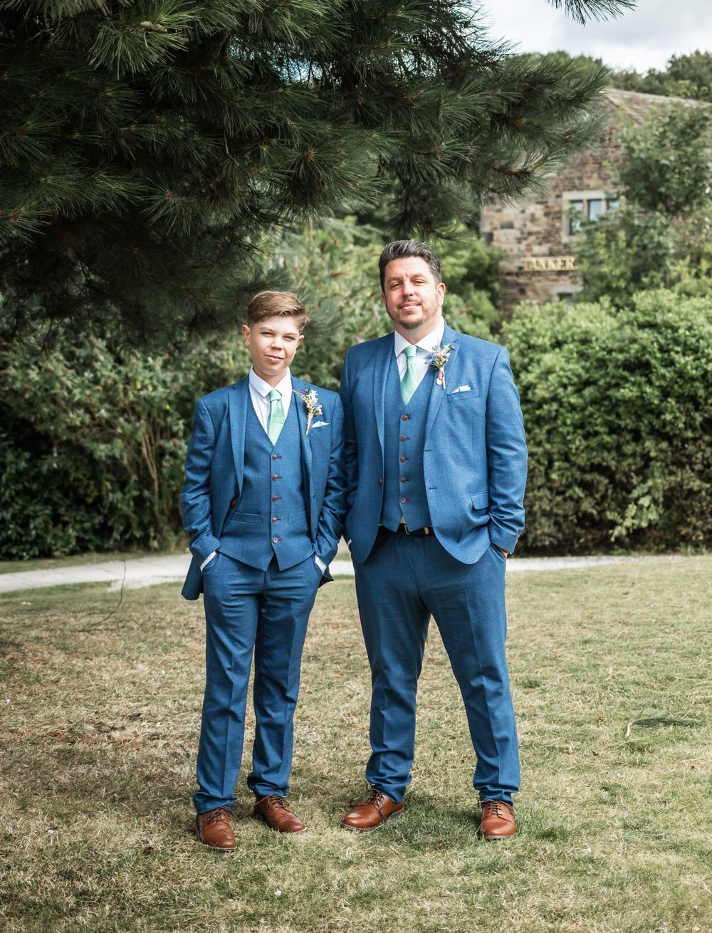 Two people in coordinated blue suits stand together on lawn with greenery and stone building behind them.