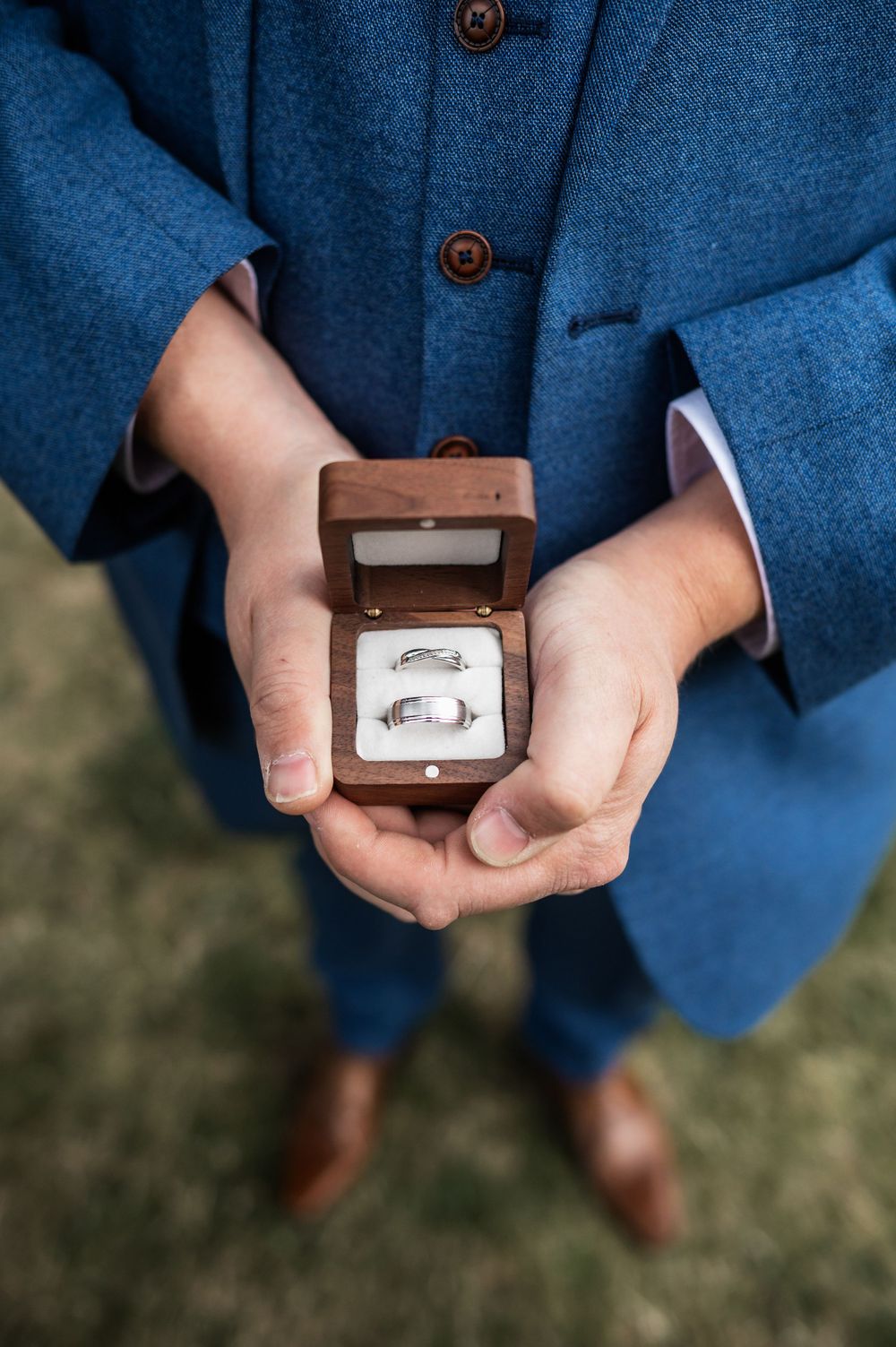 Hands holding small wooden box with wedding rings against blue suit background.