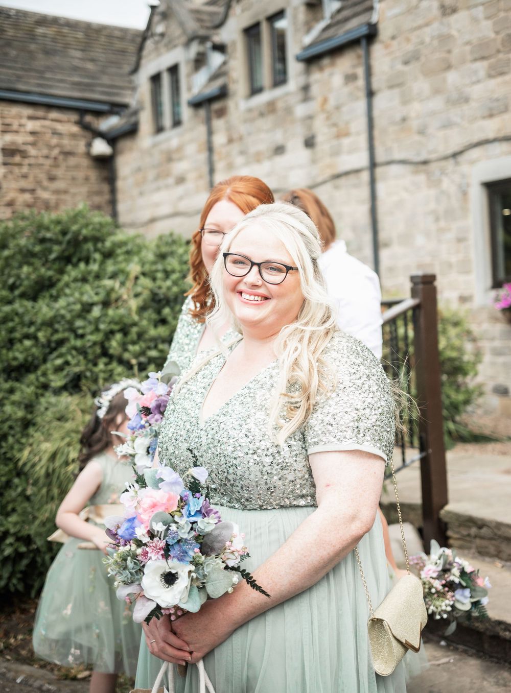 Wedding party member in sparkly mint dress holds purple and pink bouquet outside stone building.