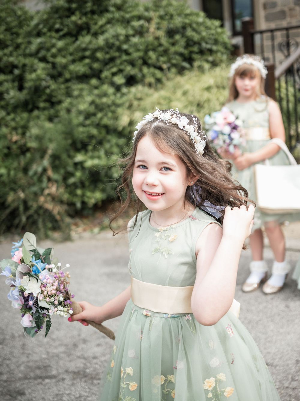 Young attendant in mint green dress with floral crown holding bouquet outdoors.