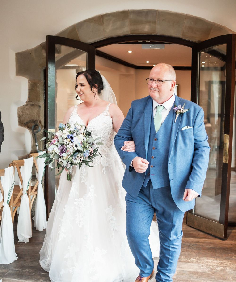 Wedding ceremony entrance with bride in white gown and companion in light blue suit.