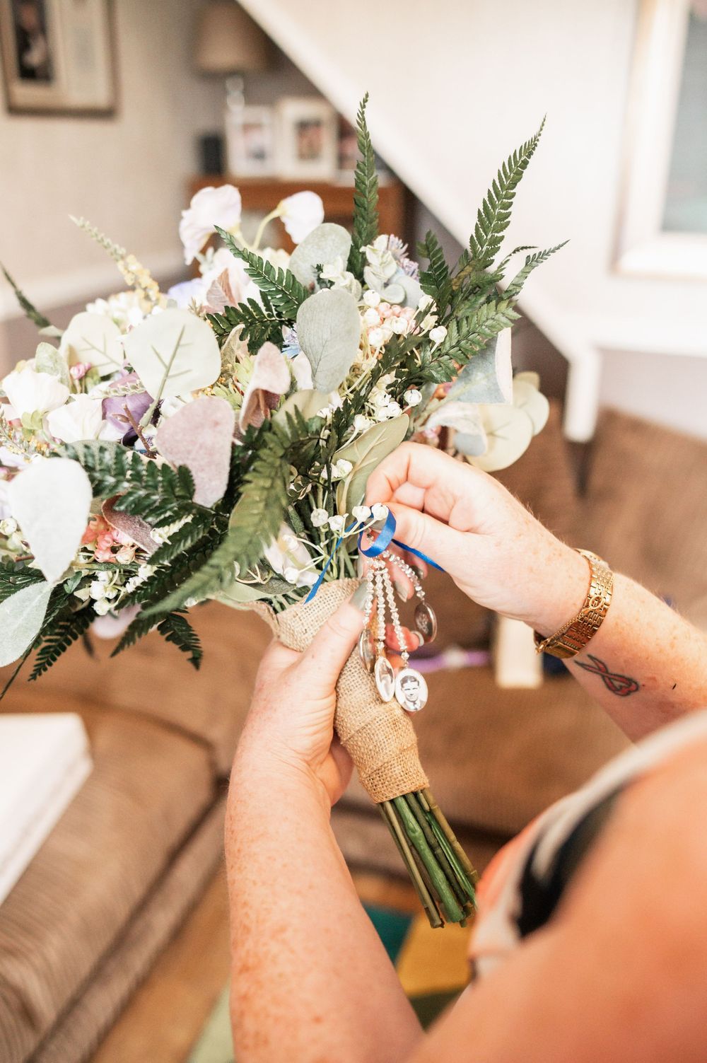 Hands adorned with jewelry arrange a delicate bouquet of white flowers and greenery against a cozy interior setting.