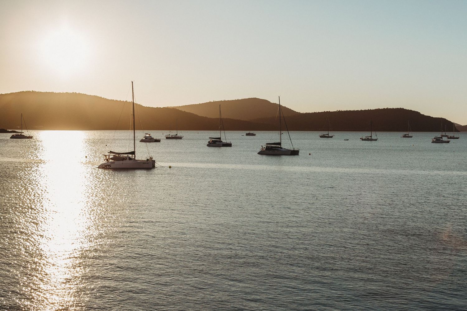 Sailboats anchored in a tranquil bay at sunset with silhouetted hills in the background.