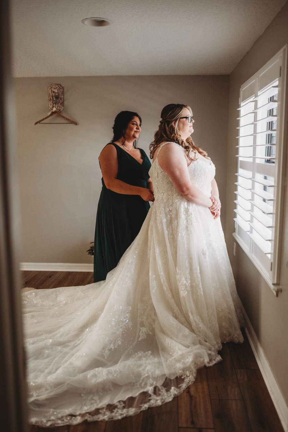 A gorgeous lace wedding gown with long train is being fitted in a room with white shuttered windows.
