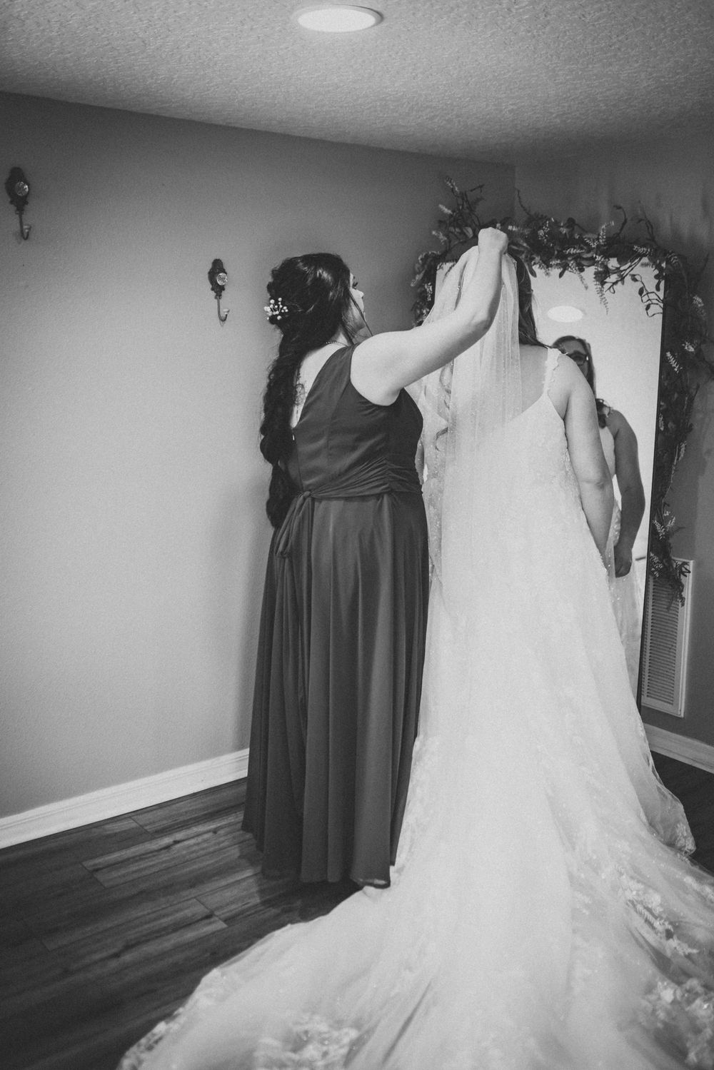 A black and white photo captures final adjustments being made to an elegant wedding dress with flowing train.