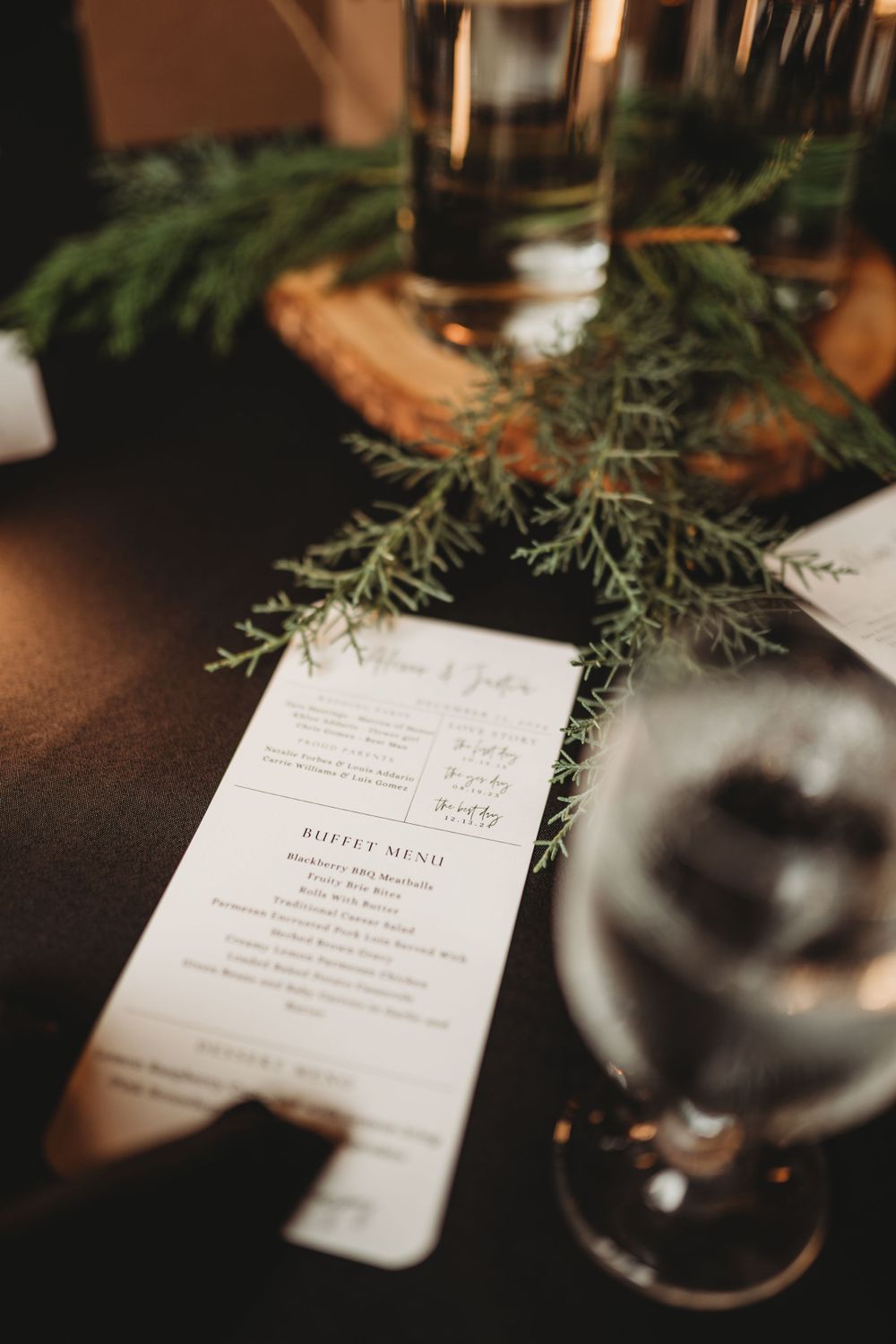 Wedding buffet menu card decorated with evergreen sprigs on a dark table setting.