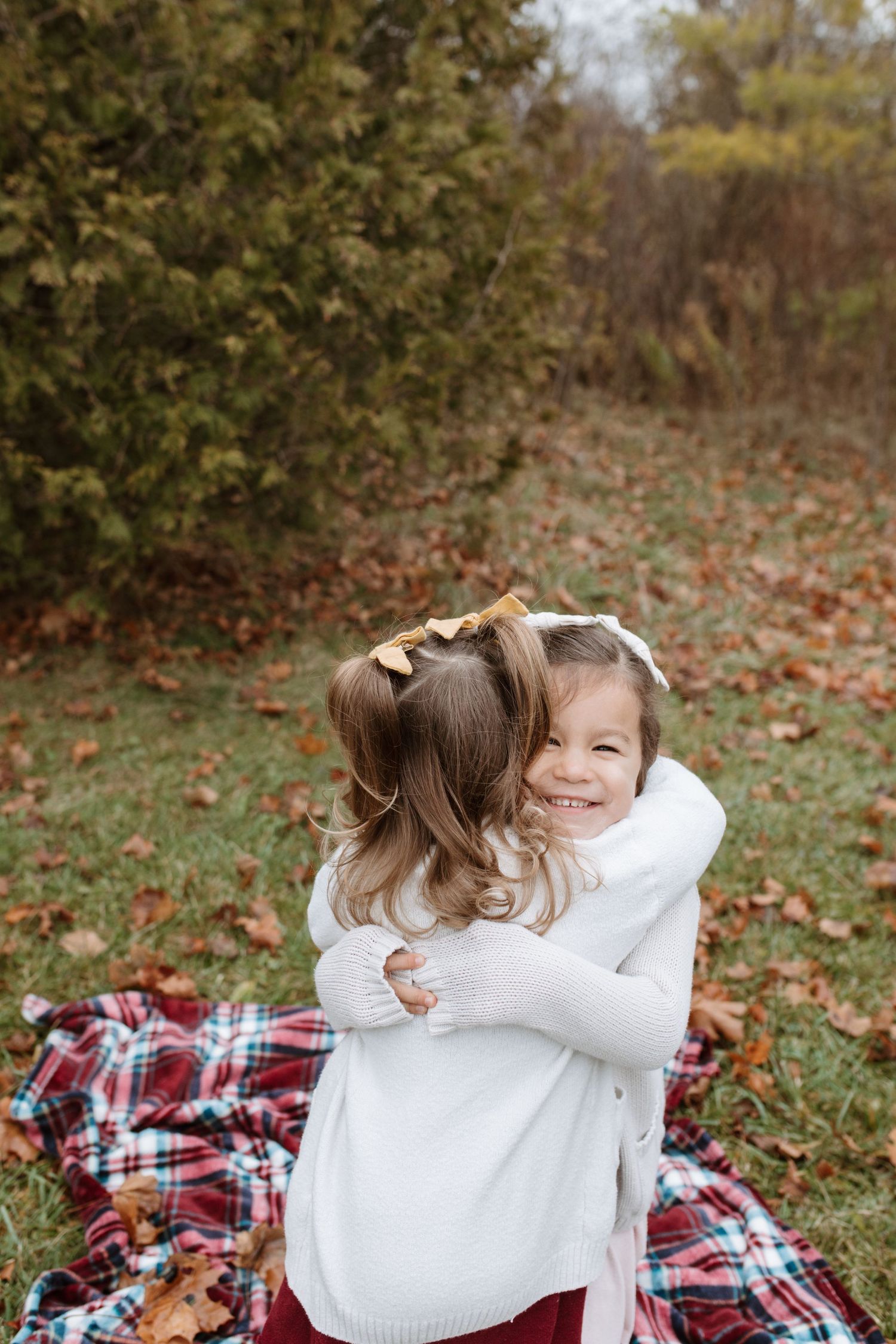 Two children wearing white sweaters embrace while sitting on a plaid blanket surrounded by fallen autumn leaves.