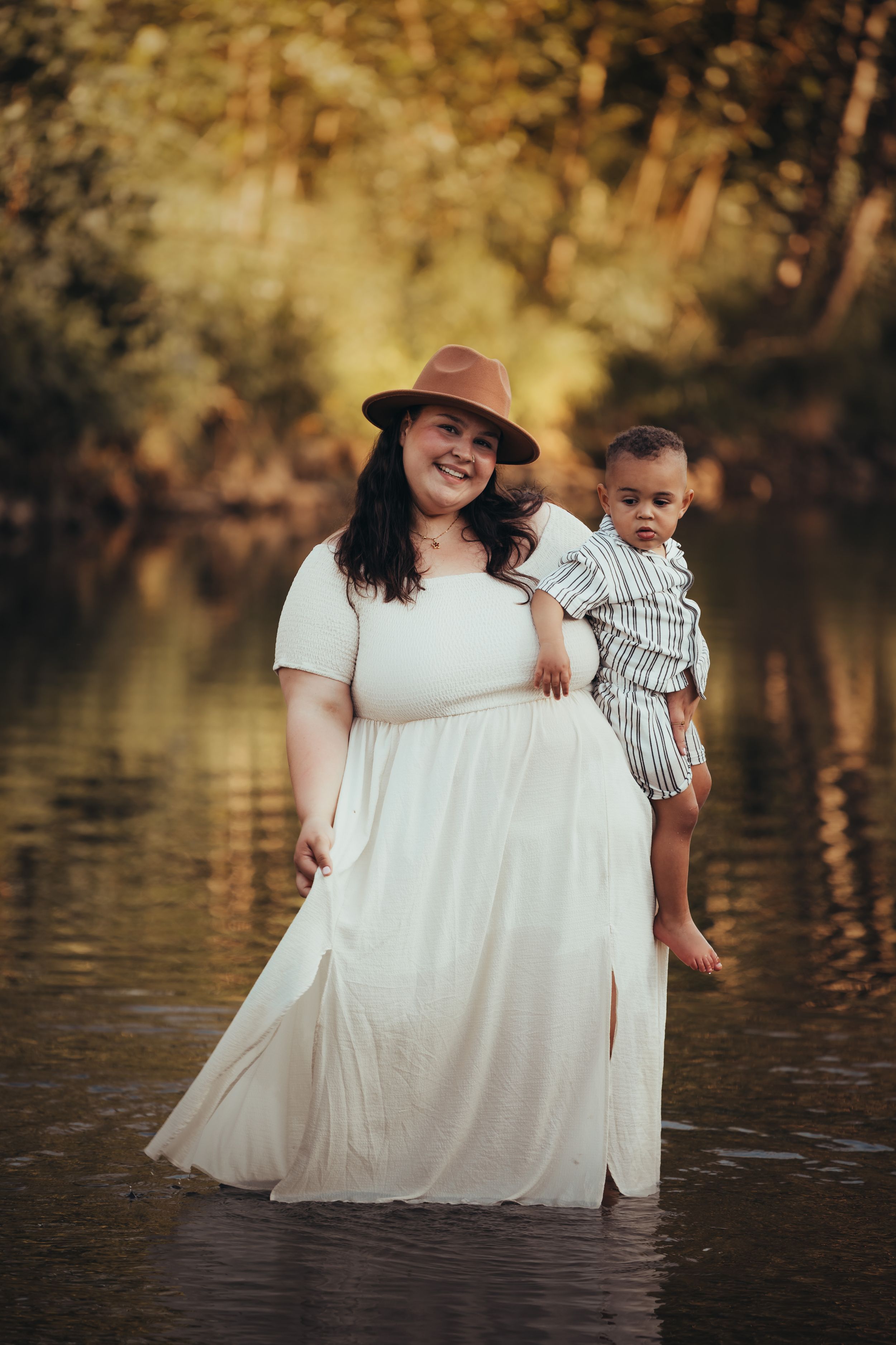 Woman in white dress and brown hat stands in water holding a small child in striped outfit at sunset.