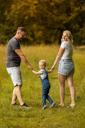 A toddler takes first steps in a grassy field while holding hands with adults.