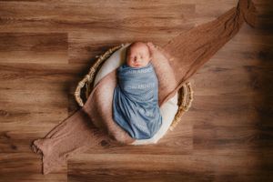 Newborn wrapped in soft blue fabric sleeps peacefully in round wooden bowl on rustic hardwood floor.