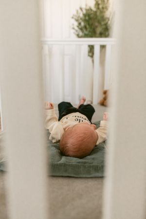 A baby lying on a green cushion viewed through white crib bars in a nursery.