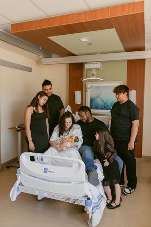 A group gathers around a hospital bed in a medical room with orange wood accents and wall art.