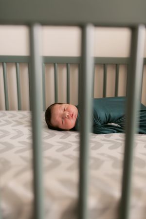 A sleeping baby rests peacefully in a crib with gray chevron bedding, viewed through white crib bars.