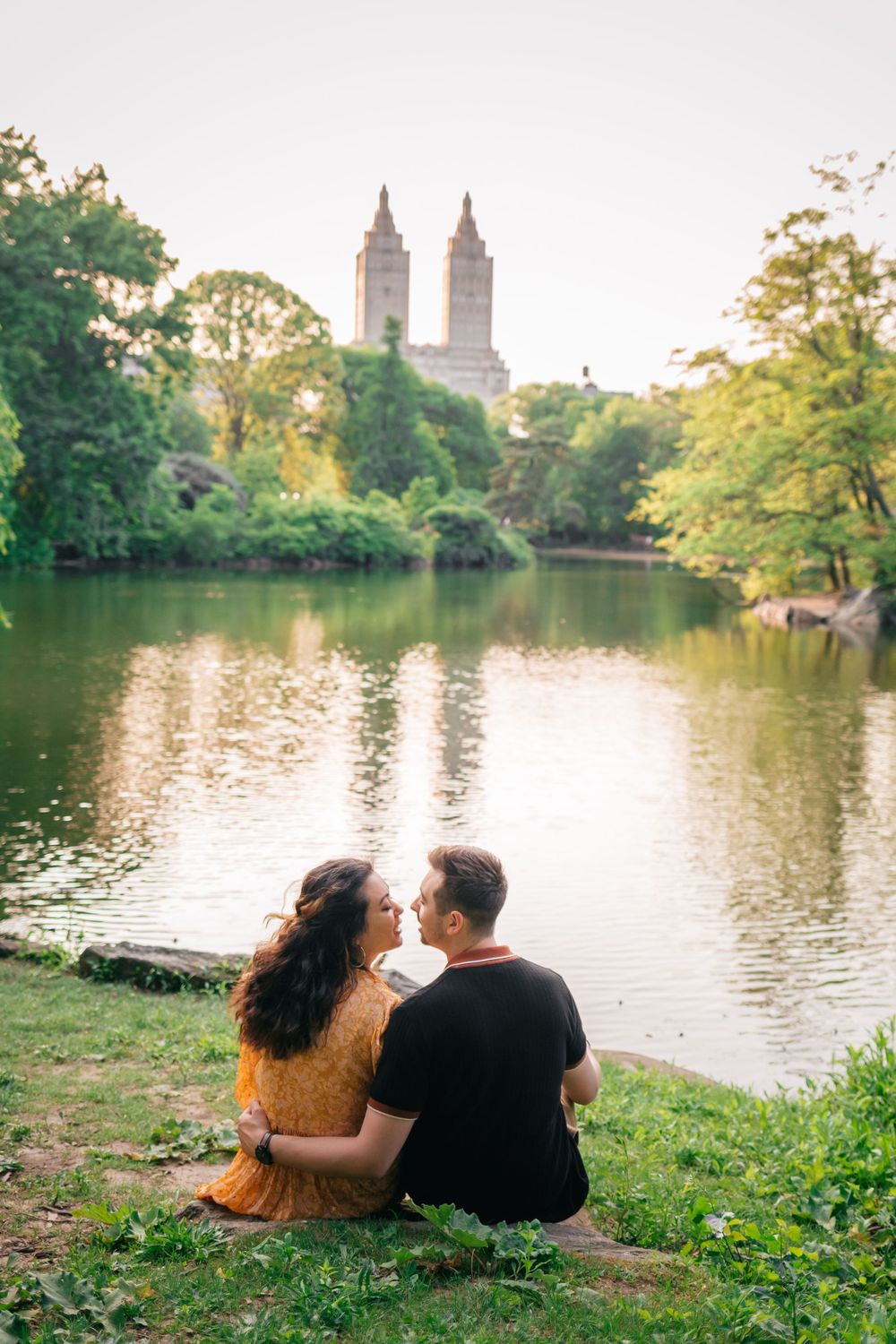 A couple sits by the lake in Central Park at sunset with iconic twin towers of the San Remo building visible in background.