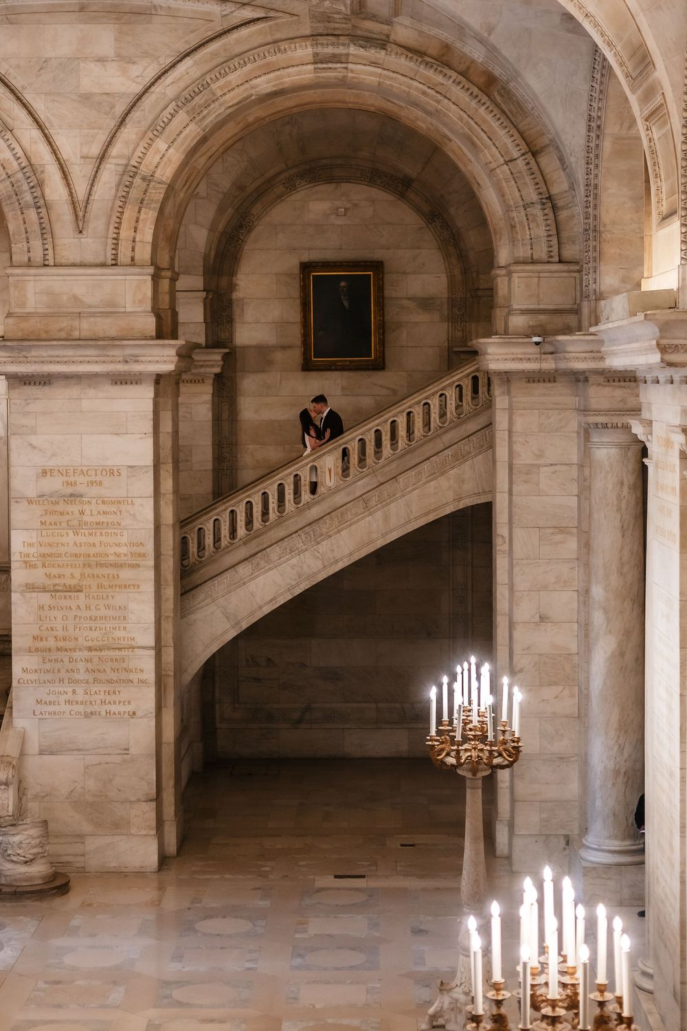 Grand marble staircase with candelabra in an ornate neoclassical library interior featuring arched ceilings and portrait painting.
